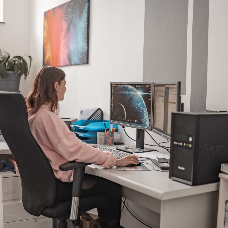 Junge Frau sitzt an einem Schreibtisch mit zwei Monitoren in einem modernen Büro.
