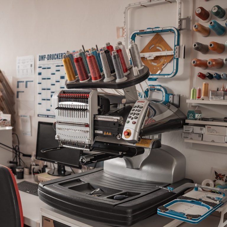 Eine Stickmaschine mit bunten Garnen auf einem Tisch, umgeben von Werkzeugen und Materialien.